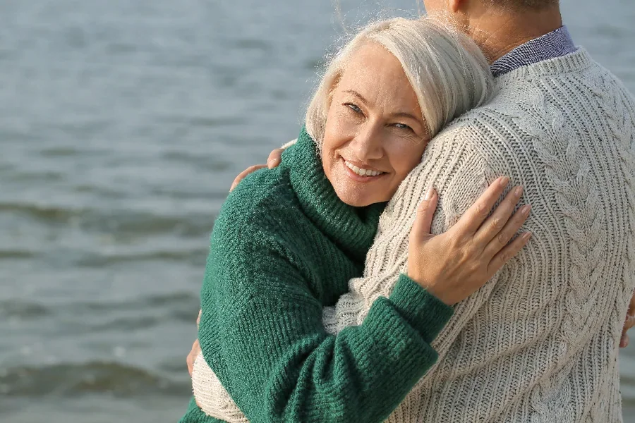 An older woman smiling while hugging her partner by the sea